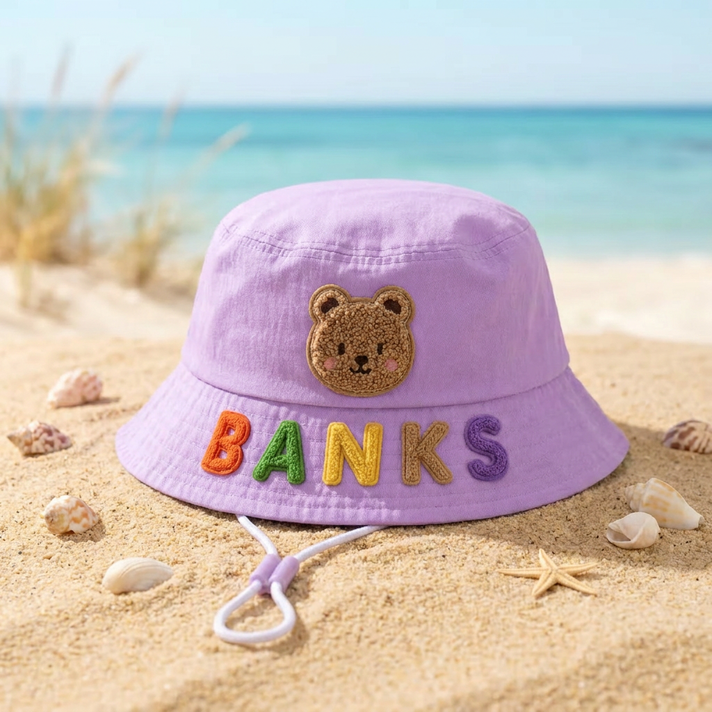 Personalized lavender kids bucket sun hat with teddy bear patch on a sunny beach